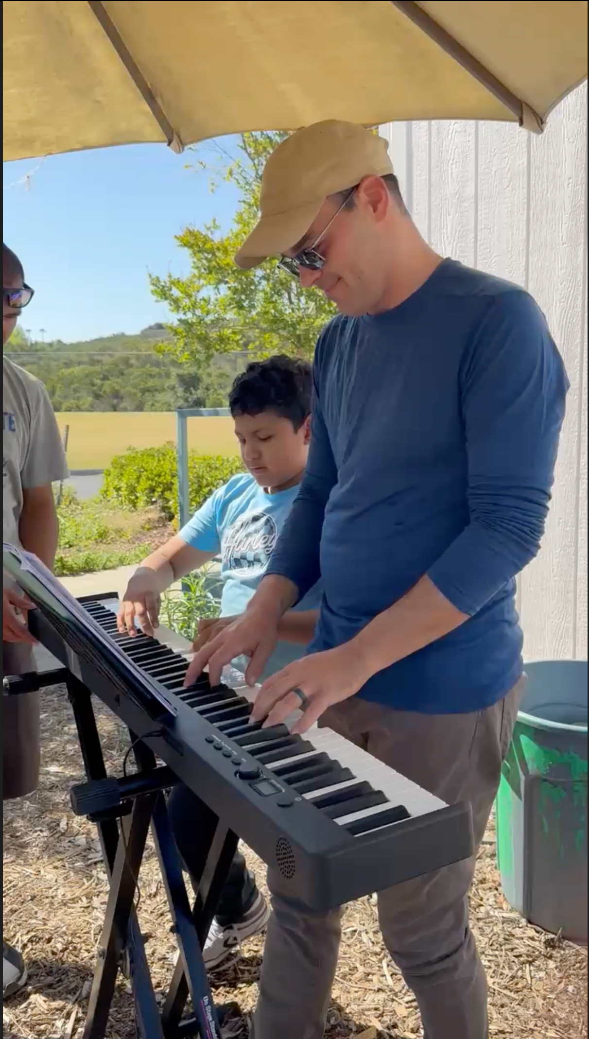 Photo of Dr. Logan Chopyk and an elementary-aged child with autism improvising on an electric keyboard at a school garden with grass and a tree in the background. Adaptive music lessons.
